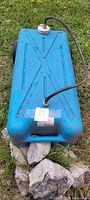 Top view showing rubber hose attached to tank cap, blue plastic body with X pattern, and wheels underneath. Signs of rust and dirt visible.