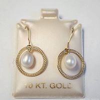 Front view of pair of 10K yellow gold earrings with oval fresh water pearls inside textured circular hoops placed on white display with '10 KT. GOLD' inscription.