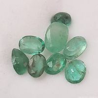 Close-up view of eight small faceted emerald gemstones on white background showing color and inclusions.