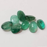 Eight loose oval faceted emerald gemstones displayed on a white background. The stones vary in shades of green, transparency, and size.