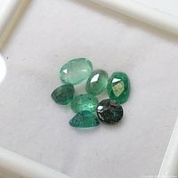Top view of seven loose emerald gemstones on white background, showing varied shapes and green hues.