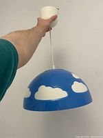 Photo of hand holding blue Ikea SKOJIG pendant lamp with white cloud pattern showing full shade and white ceiling mount.