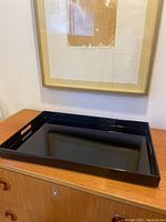 Black lacquer rectangular serving tray on wood dresser beneath framed artwork, showing top view and side handles.