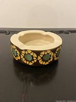 Front angle view of the round ceramic ashtray showing the floral decoration in turquoise and yellow on a dark brown base and cream interior with notches.