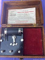Top-down view of the antique White Cross medical battery inside an open wooden box showing the black metal base, cylindrical battery component, adjustment knobs, and instruction sheet attached to the lid.