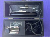 Black rectangular antique Violet Wand device with metal control panel, knob, labeled plaque, attached fabric-wrapped cord, and three glass electrode attachments stored inside case