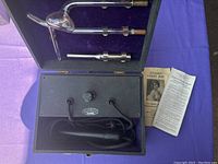 Front view of the open black box device with attached power cables, central dial, and three glass electrodes inside purple velvet lining, alongside instruction pamphlets.