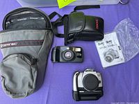 Overview of the lot showing the Tamarac camera case, compact point-and-shoot camera, Canon branded pouch, the EOS Rebel Ti camera with battery pack, and the instruction manual.