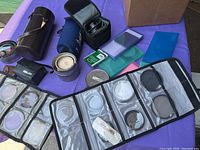 Photo showing brown leather lens case, black neoprene lens pouch, clear plastic filter wallet with filters, circular metal case, and various plastic cases and filters spread on table.