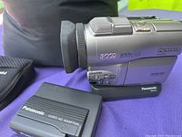 Panasonic NV-DJ100 Digicam video camera on base with Panasonic video AC adapter to the side, showing the side view with controls and branding.