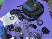 Displays Tamrac camera bag, multiple black lens covers, accessory bags, flash units, coiled cable, and various camera accessories on purple cloth.