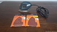 Black & Decker Mouse sander displayed with two unopened packs of sandpaper sheets suited for the sander.