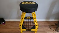 Full view of DEWALT padded swivel stool showing yellow steel frame, round padded seat with DEWALT logo, and hardwood floor background.