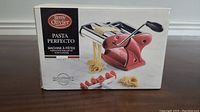Photo of the front of the box showing the Remy Olivier Pasta Perfecto pasta maker with red side plates and chrome rollers with a hand crank and pasta drying rack accessories.