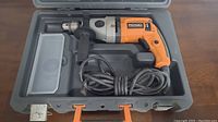 RIDGID corded hammer drill placed inside molded plastic carrying case with auxiliary handle and power cord coiled below the drill.