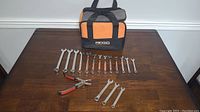 Photo showing RIDGID orange and black tool bag with 15 combination wrenches arranged by size and 1 pair of pliers on wooden table