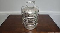 Full view of the 4-tier aluminum lunch box tiffin showing stacked cylindrical containers with engraved floral patterns and metal carrying handle