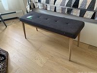 Side view of rectangular upholstered bench with tufted dark gray fabric seat and four natural wood tapered legs on hardwood floor, positioned beside a bed with quilt.