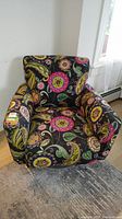 Front view of the upholstered swivel chair showing colorful floral and paisley fabric on black background, cushioned armrests and seat.
