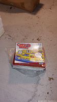 Photo showing sealed Guess Who? board game box placed on a concrete floor, with plastic wrap intact.