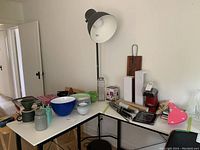 Floor lamp shown leaning against a white wall beside various kitchen and household items on folding tables.