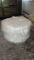 Side view of the round ottoman covered in white faux fur on a patterned carpet, showing overall shape and size.