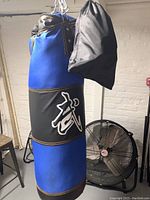 Blue and black punching bag suspended from the ceiling, with black bag hanging on the side and a fan visible in the background.