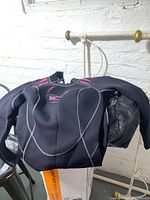 Bare brand black 3mm wetsuit laid over mesh bag with pink trim, wetsuit size large visible, hanging on metal rail against white brick wall.