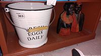 Photo of white metal bucket with black text reading 'FRESH EGGS DAILY' sitting on a shelf beside two African style statuettes.