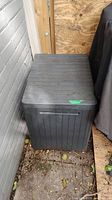 Photo of dark gray resin storage box with flat lid and vertical panel design, located outside next to wall with dirt and debris on ground.