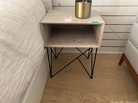Side table next to bed showing light wood top and black metal legs with geometric design.