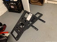 Photo of black metal swiveling TV mount with adjustable arms laid on a basement floor, showing mounting holes and swivel mechanism.
