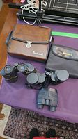 Photo of all three binoculars laid out on purple cloth including one brown and one black Bushnell case