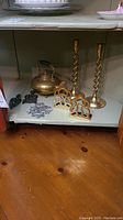Photo of two brass spiral candle holders, brass kettle, brass bookends and three metal trivets on a shelf.
