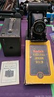 Side-by-side view of Kodak Brownie box camera with leather strap handle, Kodak Vigilant Junior Six-20 camera, its original box, and instruction booklet.