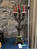 Full view of the metal candelabra standing on a blue surface near a stone wall, showing five red candles placed with white bobeches and green stone base.