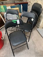 Eight black metal folding chairs with padded black vinyl seats and backs. Chairs folded and stacked in corner.
