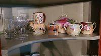 Shelf with various crystal stemware and assorted china items including pitchers, creamer, decorative items. Condition issues visible including chip and crazing.