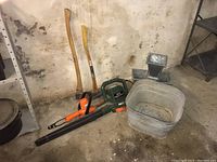 Two axes standing against wall, green blower/mulcher on floor, galvanized tub and planters to right