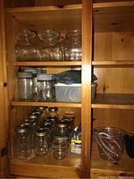 Wide shelf view showing multiple glass Mason jars, metal lids, glass canisters, and OXO measuring cups on a wooden cabinet shelf