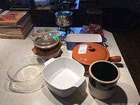 Photo showing assortment of kitchenware including Corning Ware bowls, orange fondue pot with lid, crockery pieces, and various ceramic pots and baking dishes.