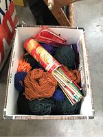 Top view of a box filled with assorted yarn skeins in various colors, a container of knitting needles, and a red pattern booklet.