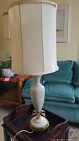Full view of the lamp with white ceramic body, gold-tone metal accents at the base, placed on a wooden surface near a blue couch.