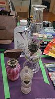 Clear glass oil lamp with floral-heart decorated base, bottle of lamp oil, pink glass lamp part, small clear glass component, metal bird decoration all on a table.