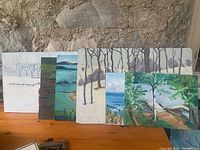 Six paintings grouped together leaning against a stone wall, showing variety of scenic landscape subjects including winter trees and lake scenes with boats and docks.
