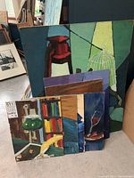 Photo showing multiple stacked oil paintings on canvas and board, displaying diverse still life subjects such as a birdcage, chair, and abstract shapes in bold colors.