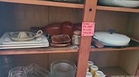 Wide shot of cabinet shelf showing stacks of white ceramic plates, brown wooden serving dish, ceramic citrus juicer, small brown dish, glass containers, and ceramic warming dish at right