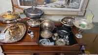 Overview of silver-plated serving dishes, Birks chafing dish, trays, and flower epergne on wooden furniture surface near window.