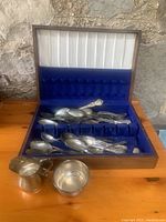Overall view of wooden box containing a collection of silver plate cutlery with creamer and sugar container in front.