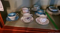 Wide view of six bone china teacup and saucer sets arranged on shelf, showing variety in colors and floral patterns, with some cups having gold trim on handles and rims.